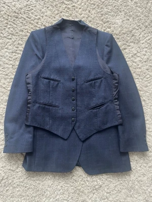 Mörkblå rutig kostym med väst - Säljer en klassisk mörkblå rutig kostym med matchande väst. Kavajen har två knappar, breda slag och fickor fram. Västen har sex knappar och fyra fickor. Perfekt för dig som vill ha en stilren och tidlös look.