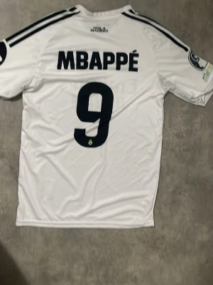 Real Madrid matchtröja Mbappé 9 - Säljer en vit Real Madrid matchtröja från Adidas med Mbappé och nummer 9 på ryggen. Tröjan har svarta detaljer, klubbmärke på bröstet och sponsortryck 'Emirates Fly Better' framtill. Tillverkad i ett lätt och ventilerande material, perfekt för fotbollsträning eller match.