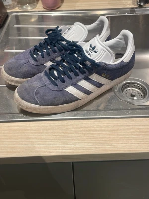 Adidas Gazelle  - Snygga Adidas Gazelle sneakers i blå mocka med vita detaljer och klassiska tre ränder på sidorna. Skorna har mörkblå snören, vit sula och vit plös med Adidas-logga. Perfekta för en stilren och sportig look. Skorna är använda men inga defekter osv, fler frågor eller funderingar så hör av er i dm!