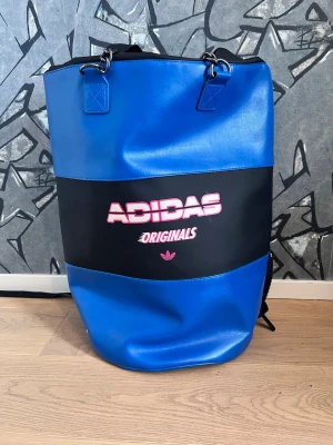 Blå Adidas Originals shoppingväska - Snygg ryggsäck från Adidas Originals i blått och svart konstläder med stor logga i vitt och rosa framtill. Väskan har kedjehandtag i metall och rymlig cylinderformad design, perfekt för att bära med sig allt man behöver. Cool streetstyle-vibe och praktisk för vardagsbruk.