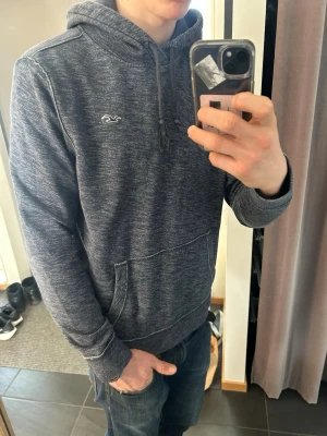 Mörkblå hoodie från Hollister - Säljer en mörkblå hoodie från Hollister med melerad look och klassisk logga på bröstet. Hoodien har huva med dragsko, känguruficka framtill och ribbade muddar vid ärmar och nederkant. Perfekt för en avslappnad stil.