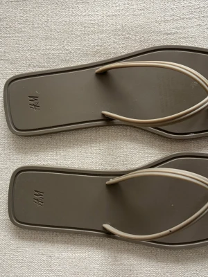 Bruna flip-flops från H&M - Stilrena bruna flip-flops från H&M med fyrkantig form och tunna, ljusbeige remmar. Perfekta för sommarens alla äventyr. Aldrig använda 