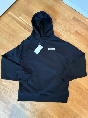 Jacquemus Hoodie Helt Ny!!! - Svart hoodie från Jacquemus med minimalistisk design och liten vit Jacquemus-logga på bröstet. Klassisk passform med huva och ribbade muddar. Perfekt för dig som gillar stilrena och trendiga plagg. Materialet känns mjukt och bekvämt.
