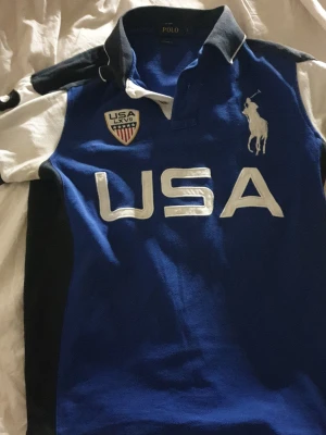 Blå Polo Ralph Lauren USA t-shirt - Snygg blå t-shirt från Polo Ralph Lauren med vit USA-text och broderad logga på bröstet. T-shirten har krage, korta ärmar och detaljer i vitt och svart. Perfekt för dig som gillar sportig och klassisk stil. Storleken är mer M,S. Litet litet hål i sidan kolla sista bilden märks knappt