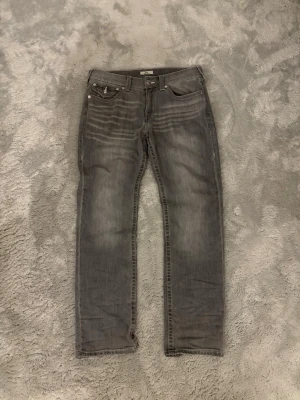 Grå jeans från True Religion - Säljer ett par grå jeans från True Religion med raka ben och klassiska kontrastsömmar. Jeansen har fem fickor, snygga detaljer på bakfickorna och en patch med logga bak i midjan. Perfekta för en avslappnad och trendig look