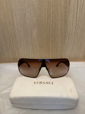 Versace solglasögon  - Exklusiva Versace solglasögon i shield-design med ikonisk Medusa-detalj på skalmarna. Tidlös statement-modell med brun tonad lins och lyxiga gulddetaljer.  • Original Versace • Mycket fint skick • Original Versace-fodral ingår • Bekväma och lätta • Trendig Y2K / luxury-look  Nypris ca 3 500–4 000 kr. Säljs då de inte används.  Skickas snabbt
