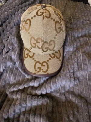 Beige keps från Gucci med GG-mönster - Säljer en beige keps från Gucci med det klassiska GG-monogrammet i brunt över hela kepsen. Kepsen har böjd skärm med mörkbrun kant och är tillverkad i canvasmaterial. Perfekt för dig som gillar exklusiva accessoarer och vill sticka ut.