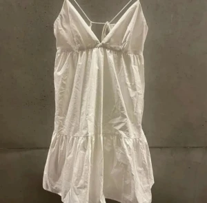 Vit kortklänning med volang - Supersöt vit kortklänning med tunna axelband och v-ringad urringning. Klänningen har en volang nedtill och är tillverkad i ett lätt bomullstyg som ger en luftig känsla. Perfekt för varma dagar och har en feminin, flowy siluett.