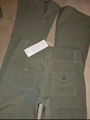 Gröna bootcut byxor med fickor - Snygga gröna bootcut byxor med två bakfickor som har lock och knapp. Byxorna har breda benslut och markerade sömmar. Perfekta för dig som gillar en retroinspirerad look och vill ha något bekvämt men ändå stilrent.