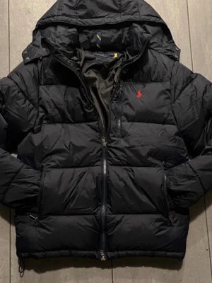 Svart pufferjacka från Polo Ralph Lauren - Snygg svart pufferjacka från Polo Ralph Lauren med röd logga på bröstet. Jackan har huva, dragkedja framtill, två sidofickor med dragkedja och justerbar dragsko nedtill. Perfekt för kalla vinterdagar och riktigt skön att bära. Justeringen vid höften är sönder men funkar endå och på dragkedjan är två tänder borta men funkar och stänga endå