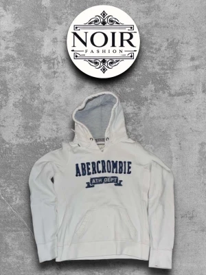 Vit hoodie från Abercrombie - Vit hoodie från Abercrombie med blå broderad logga och text på bröstet. Klassisk känguruficka framtill och huva med ljusgrått foder. Perfekt för en avslappnad och sportig stil.