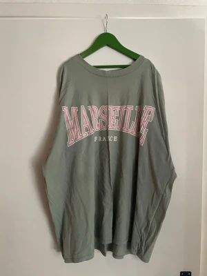 Grön t-shirt med Marseille-tryck - Cool grön t-shirt med stort Marseille-tryck i rosa och vitt på bröstet. Skön passform och rund halsringning, perfekt för dig som gillar streetstyle och franska vibes. Materialet känns mjukt och bekvämt, perfekt för chill dagar.