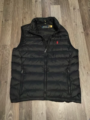 Svart dunväst från Polo Ralph Lauren - Snygg svart dunväst från Polo Ralph Lauren med klassisk quiltad design och röd logga på bröstet. Västen har hög krage, dragkedja framtill och två fickor med dragkedja. Perfekt för lager-på-lager och håller dig varm under kyliga dagar. Passar s
