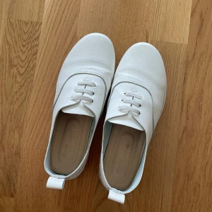 Vita sneakers från Bata - Snygga vita slop-ons med klassisk design och rund tå. Skorna har dekorativ snörning framtill och är gjorda i ett slätt syntetmaterial med diskret struktur. Platt sula och dragflik bak för enkel påtagning. Perfekta för en clean och stilren look.