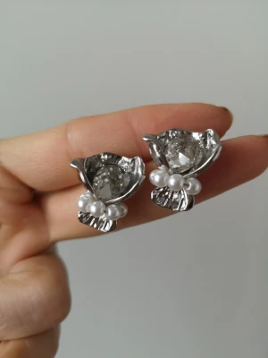 Silvriga örhängen med pärlor och strass - Unika örhängen i silverfärgad metall med en stor klar strass i mitten och små vita pärlor som dekoration. Blomformad design som ger en cool och elegant look. Perfekta för dig som vill sticka ut med accessoarerna.