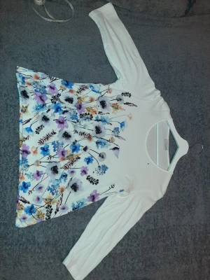 Vit blommig topp med trekvartsärm från Cotswold Collections  - Snygg vit topp med färgglatt blommigt mönster i lila, blått, svart och orange. Modellen har rund halsringning och trekvartsärm. Perfekt för dig som gillar en feminin och fräsch stil. Materialet känns mjukt och bekvämt.