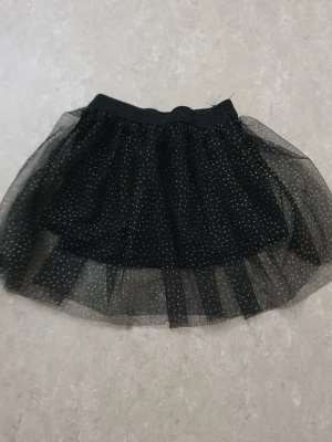 Kjol - Säljer en svart kjol med glittriga prickar och ett lager av transparent tyll. Kjolen har elastisk midja och ger en cool och lekfull look. Perfekt för dig som vill sticka ut med lite extra glitter.