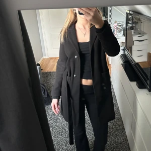 Svart kappa med knytskärp - Stilren svart lång kappa med knytskärp. Klassisk rak passform som passar till många olika outfits. Perfekt för dig som vill ha en enkel men snygg look. 
