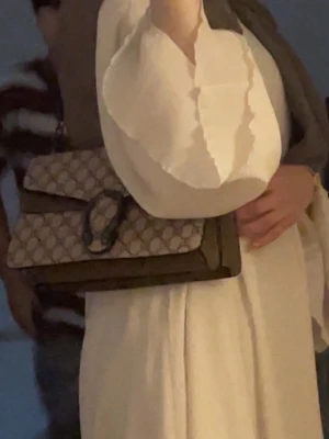 Gucci Dionysus beige axelväska - Säljer en Gucci Dionysus axelväska i beige canvas med GG-mönster och detaljer i mocka. Väskan har en silverfärgad kedjerem och ett snyggt spänne framtill. Perfekt för dig som vill ha en ikonisk och trendig väska med lyxig känsla.