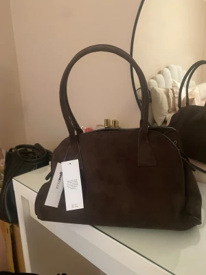 Mörkbrun mockaväska från Gina Tricot - Helt ny snygg handväska i mörkbrun mocka från Gina. Väskan har rundad form, två rejäla handtag och två rosetter som knytning på sidorna. Perfekt storlek för det viktigaste och ger en stilren look till din outfit.