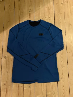 Helly Hansen underställ - Snygg blå långärmad funktions-tröja från Helly Hansen med svarta detaljer och HH-logga på bröstet. Tillverkad i ett tekniskt material som andas, perfekt för träning eller långa vandringar. Sprillans ny kostar Ca 600kr. Tveka inte på att höra av dig :)