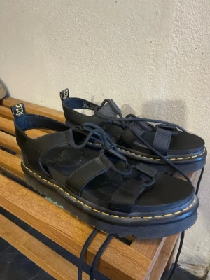 Svarta Dr. Martens sandaler i läder - Chunky svarta sandaler från Dr. Martens i äkta läder med klassisk gul söm runt sulan. Sandalerna har flera remmar, justerbar snörning och grov platåsula. Ikoniska dragflikar bak med gul text. Perfekta för en edgy och trendig stil.