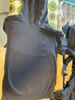 Svart hoodie med huva - En enkel svart hoodie med huva och ribbade muddar vid ärmslut och nederkant. Hoodien har en klassisk passform och är tillverkad i ett mjukt bomullsliknande material. Perfekt för en avslappnad stil.