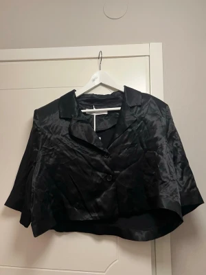 NY Svart cropped kavaj från Lis Bonne - NY Snygg svart cropped kavaj från Lis Bonne Atelier i glansigt material. Kavajen har klassisk krage, två knappar framtill och kortare passform som ger en trendig look. Perfekt att styla till både jeans och kjol för en cool outfit.