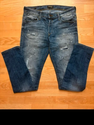 Blå slitna jeans från Jack & Jones - Säljer ett par blå jeans från Jack & Jones med snygga slitningar och lätt tvättad look. Jeansen har klassisk femficksdesign, normal midja och raka ben. Perfekta för en avslappnad och trendig stil.