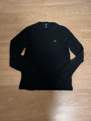 Svart långärmad tröja från Levi's - Klassisk svart långärmad tröja från Levi's med rund halsringning och liten röd Levi's-logga på bröstet. Tröjan har en enkel och stilren design, perfekt för dig som gillar basic plagg med en touch av streetwear.