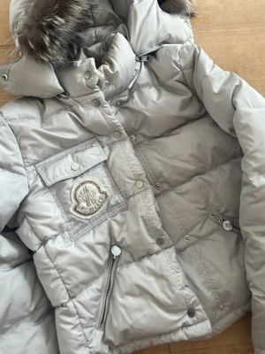 Ljusgrå dunjacka från Moncler pälskrage - Snygg ljusgrå/silver dunjacka från Moncler med stor huva och pälsdetalj. Jackan har flera fickor med dragkedja och tryckknappar samt Moncler-logga på bröstet. Perfekt för kalla vinterdagar och riktigt varm tack vare dunfyllningen.