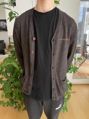 Mörkbrun kofta från Nudie Jeans - Snygg mörkbrun kofta från Nudie Jeans med V-ringning och fem knappar framtill. Koftan har två fickor och diskret broderad logga i gult på bröstet. Tillverkad i mjukt stickat material, perfekt för lager på lager och chill stil.