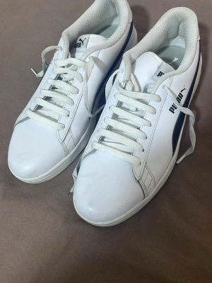 Vita Puma sneakers med svart detalj - Snygga vita sneakers från Puma med klassisk svart sidodetalj och logga på både plös och häl. Skorna har vita snören och en låg profil, tillverkade i slätt syntetmaterial med bekväm vadderad insida. Perfekta för en clean och sportig look.