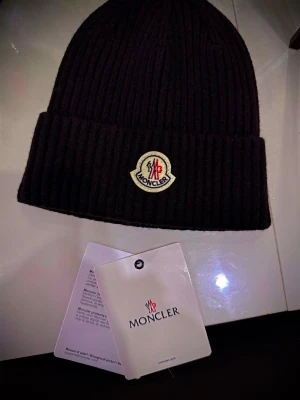 Svart Moncler mössa i ull - Säljer en svart ribbstickad mössa från Moncler med uppvikt kant och klassisk Moncler-logga framtill. Mössan är tillverkad i mjuk ull och har en stilren, tidlös design som passar perfekt till kalla dagar.