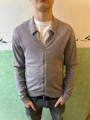 Ljusgrå ziptröja från Riley - Snygg ljusgrå ziptröja från Riley med klassisk krage och dragkedja framtill. Tröjan är tillverkad i mjukt material och har ribbade muddar vid ärmslut och nederkant. Perfekt att slänga på sig över en t-shirt för en clean look.