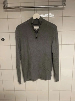 Grå stickad tröja med halv dragkedja - Snygg grå stickad tröja med halfzip och ståkrage. Tröjan har lång ärm och är tillverkad i ett mjukt, tunt stickat material. Perfekt för lager-på-lager och enkel att matcha med olika outfits.