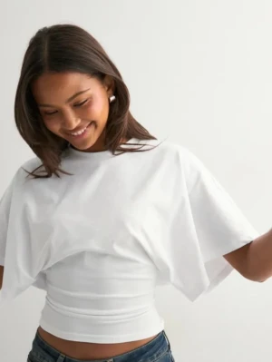 Vit croppad topp från Nelly - Säljer en vit croppad topp från Nelly med vida, korta ärmar och rund halsringning. Toppen har en loose passform upptill och sitter tightare i midjan, vilket ger en snygg siluett. Perfekt att styla med jeans eller kjol.