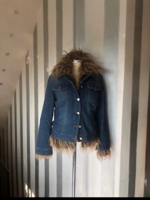 Jeansjacka med fuskpälsdetaljer - Mörkblå jeansjacka med avtagbar fuskpäls runt krage, ärmslut och nederkant. Klassisk knappstängning framtill och två bröstfickor med lock. Snygg och unik jacka som sticker ut i mängden.