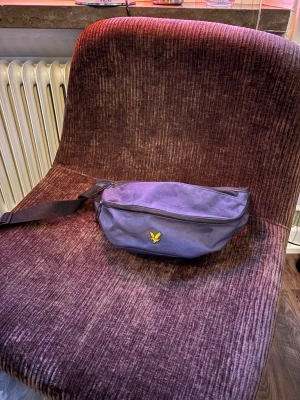 Blå magväska  från Lyle & Scott - Snygg Blå magväska från Lyle & Scott med klassisk gul logga framtill. Väskan har justerbart svart band och spänne, samt dragkedja upptill. Perfekt för att bära det viktigaste och enkel att slänga över axeln. Har en fläck ovanpå. Kan gå ner i pris!