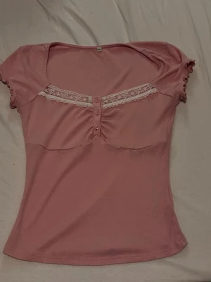 Rosa topp med spets och knappar - Söt rosa topp med korta ärmar, spetsdetalj och små knappar framtill. Toppen har en lätt rynkad effekt vid bysten och är figurnära i modellen. Perfekt för dig som gillar en feminin och trendig stil.