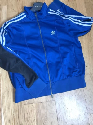 Blå Adidas track jacket med dragkedja - Snygg blå track jacket från Adidas med klassiska vita ränder längs ärmarna och Adidas-logga på bröstet. Jackan har dragkedja framtill, ståkrage och ribbade muddar. Materialet är glansigt och sportigt, perfekt för träning eller streetwear.