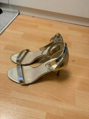 Guldiga pumps med smal rem från XIT - Snygga guldiga pumps från XIT med öppen tå, smal rem över foten och justerbart spänne vid ankeln. Klacken är smal och elegant, perfekt för att lyfta vilken outfit som helst. Tillverkade i ett glansigt syntetmaterial som ger en lyxig känsla.