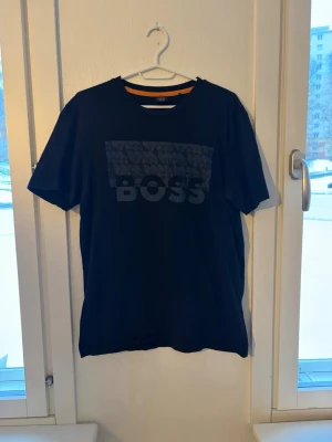 Svart BOSS t-shirt med tryck - Snygg svart t-shirt från BOSS med stort ton-i-ton-tryck på bröstet där det står 'BOSS'. Klassisk rund halsringning och korta ärmar. Perfekt för en stilren och avslappnad look.