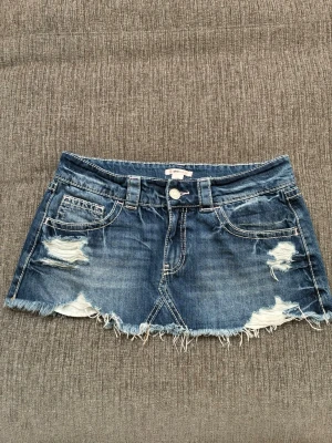 Blå kort jeanskjol med slitningar - Kort blå jeanskjol med slitna detaljer och fransig nederkant. Klassisk femficksmodell med bälteshällor och dragkedja fram. Från Gina Tricot 14+. Använd men i mycket bra skick!