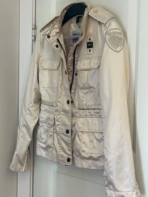 Beige jacka från Blauer med fickor - Stilren beige jacka från Blauer med flera praktiska fickor, axelklaffar och patch på ärmen. Jackan har dragkedja och knappar framtill samt en klassisk krage. Perfekt för dig som gillar en clean och funktionell look.