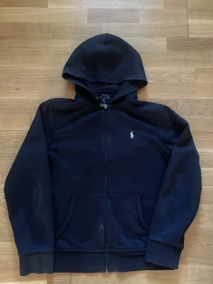 Svart hoodie från Polo Ralph Lauren - Klassisk svart hoodie med dragkedja från Polo Ralph Lauren. Har huva, två fickor framtill och den ikoniska vita loggan broderad på bröstet. Perfekt för en avslappnad och stilren look.