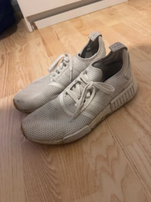 Vita Adidas NMD_R1 sneakers - Säljer ett par vita Adidas NMD_R1 sneakers med stickad ovandel i mesh och klassiska tre ränder på sidan. Skorna har en chunky sula med Boost-dämpning och detaljer i gummi. Perfekta för dig som gillar streetwear och bekväma sneakers.
