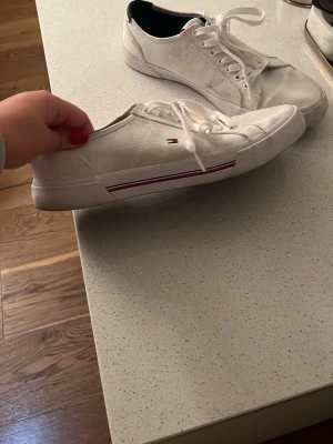 Vita sneakers från Tommy Hilfiger - Snygga vita sneakers från Tommy Hilfiger med klassisk låg profil och snörning. Skorna har diskret logga på sidan och svarta detaljer vid hälen. Tillverkade i textil med gummisula och röda samt blå ränder längs sidan för en sportig look.