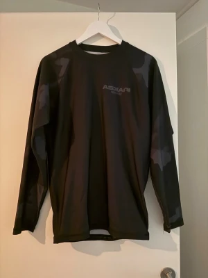 Rashguard Askari - Cool svart långärmad rash från Askari med diskret camouflagemönster på ärmar och axlar. Tillverkad i ett syntetmaterial som känns lätt och smidigt. Perfekt för dig som gillar en sportig och avslappnad stil.
