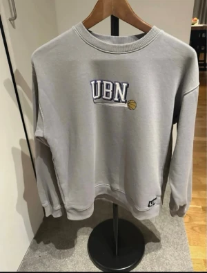 Grå sweatshirt med UBN-tryck - Säljer en grå sweatshirt från UBN med tryck i blått och vitt samt en liten basketboll på bröstet. Tröjan har rund hals, långa ärmar och ribbade muddar. Perfekt för en sportig och avslappnad stil.